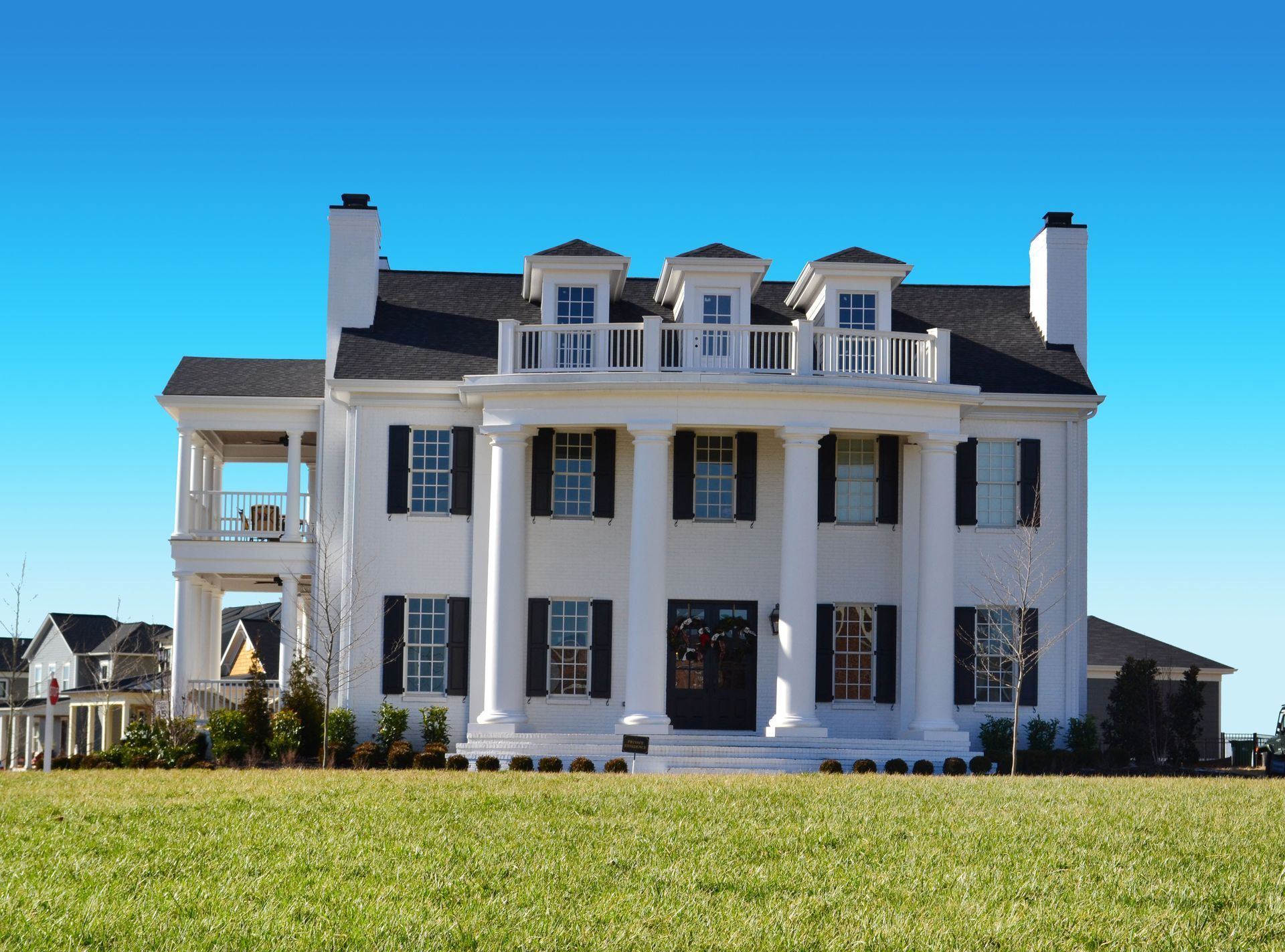 White two-story house with black shutters, large columns, and a grassy lawn under a blue sky.