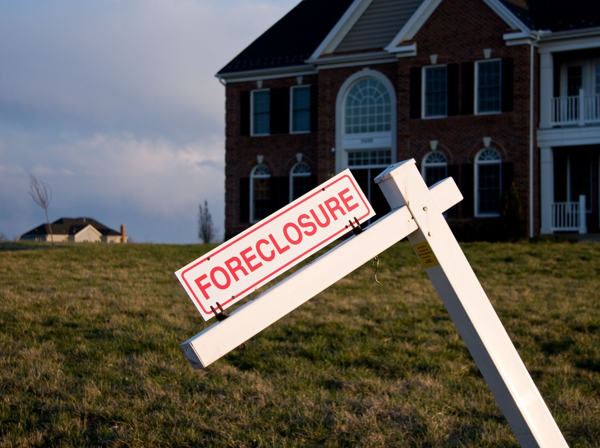 Foreclosure sign in front of a large house on a grassy lot under a cloudy sky.