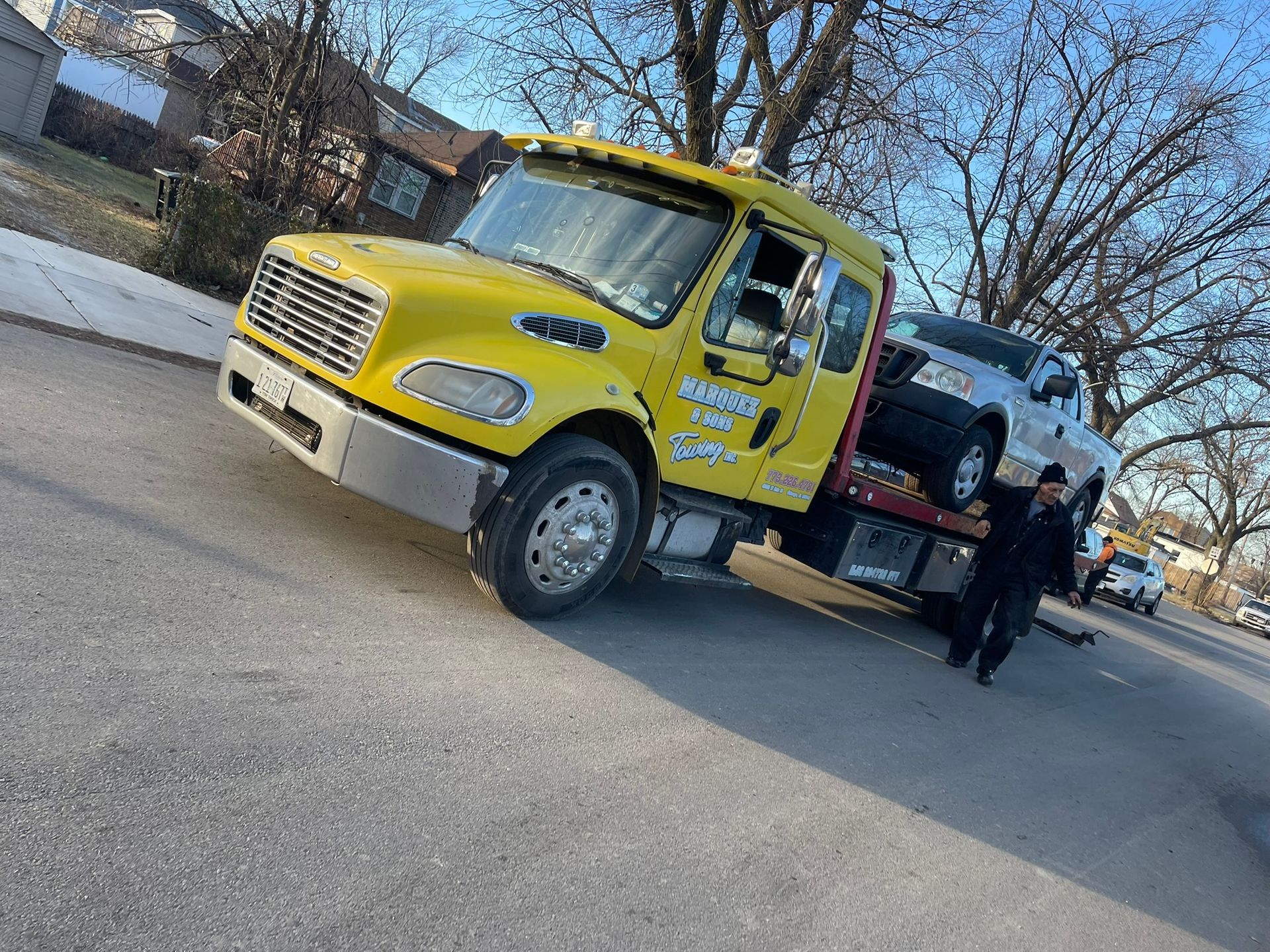 Yellow tow truck towing a car on a street. Man stands nearby.