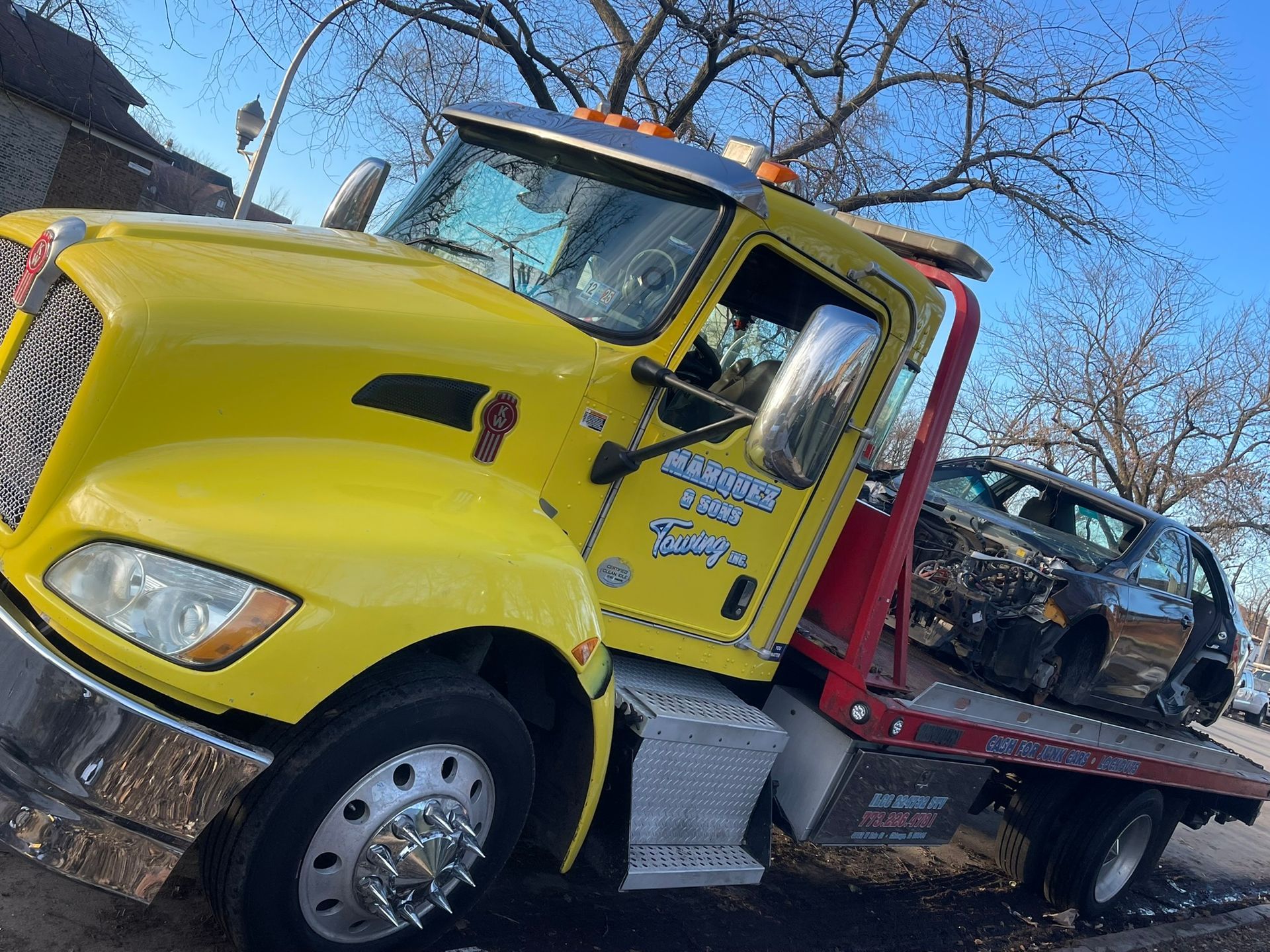Yellow tow truck carrying a damaged black car on a sunny day.