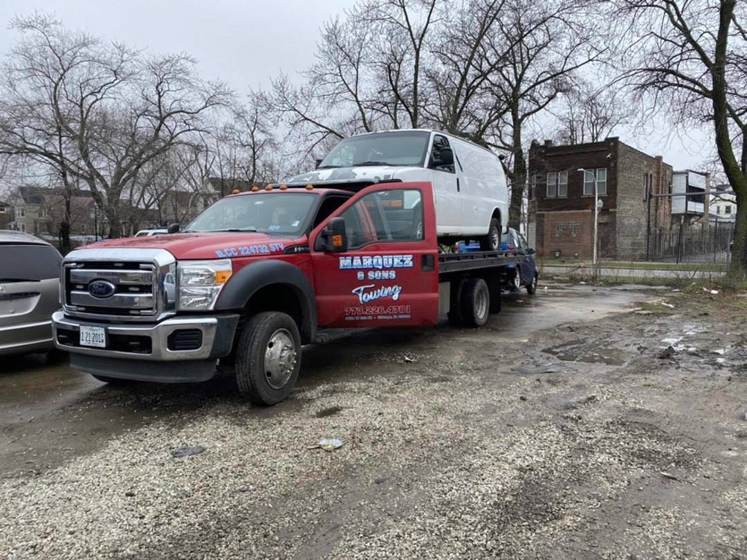 Red tow truck carrying a white van, parked on a dirt lot. Buildings and trees in background.