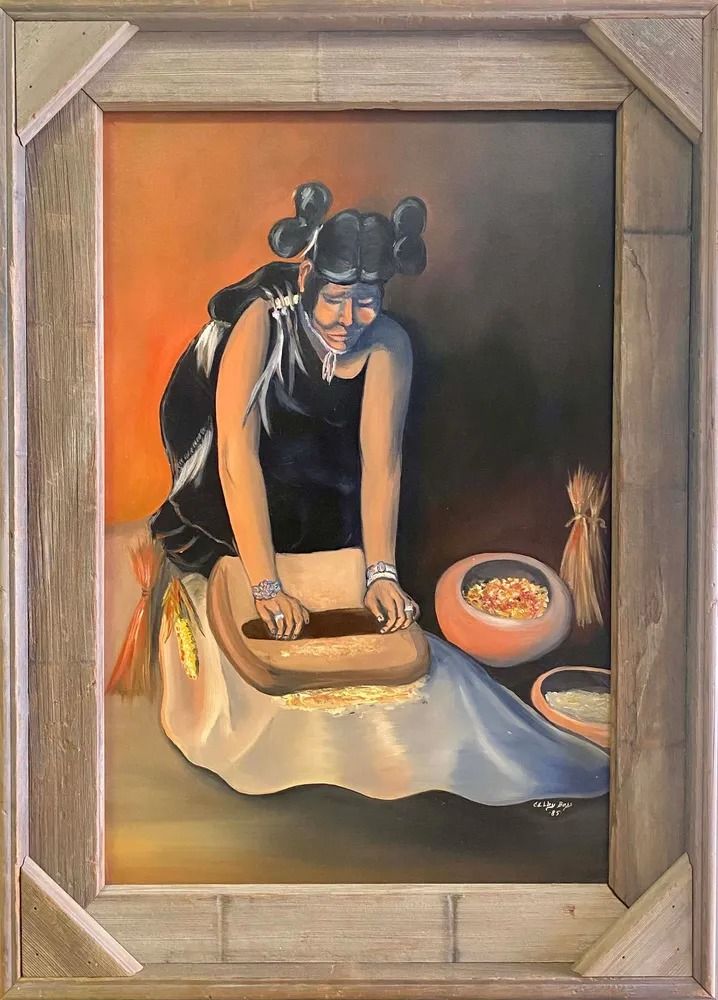 A woman in Native American dress grinds on a stone, with bowls of food, framed in wood.