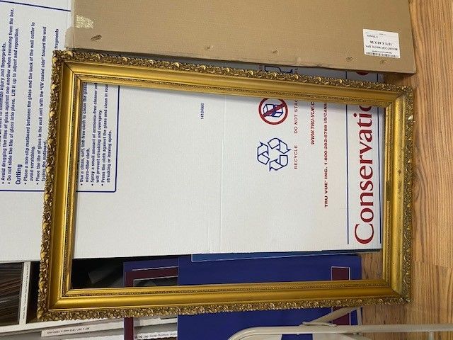 Gold ornate picture frame on top of a white cardboard box with conservation label.
