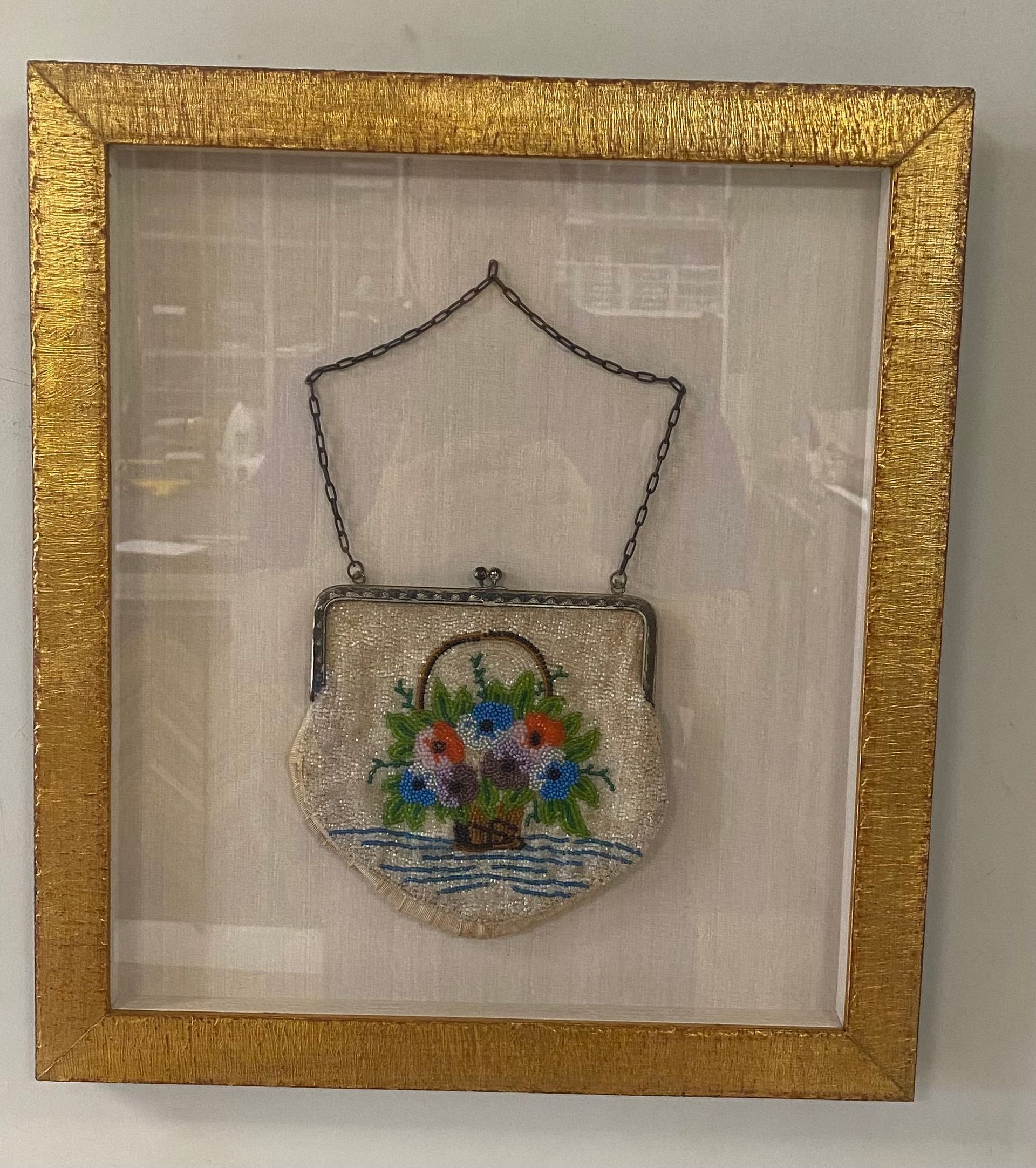Framed beaded purse with a floral basket design, in a gold frame on a textured white background.