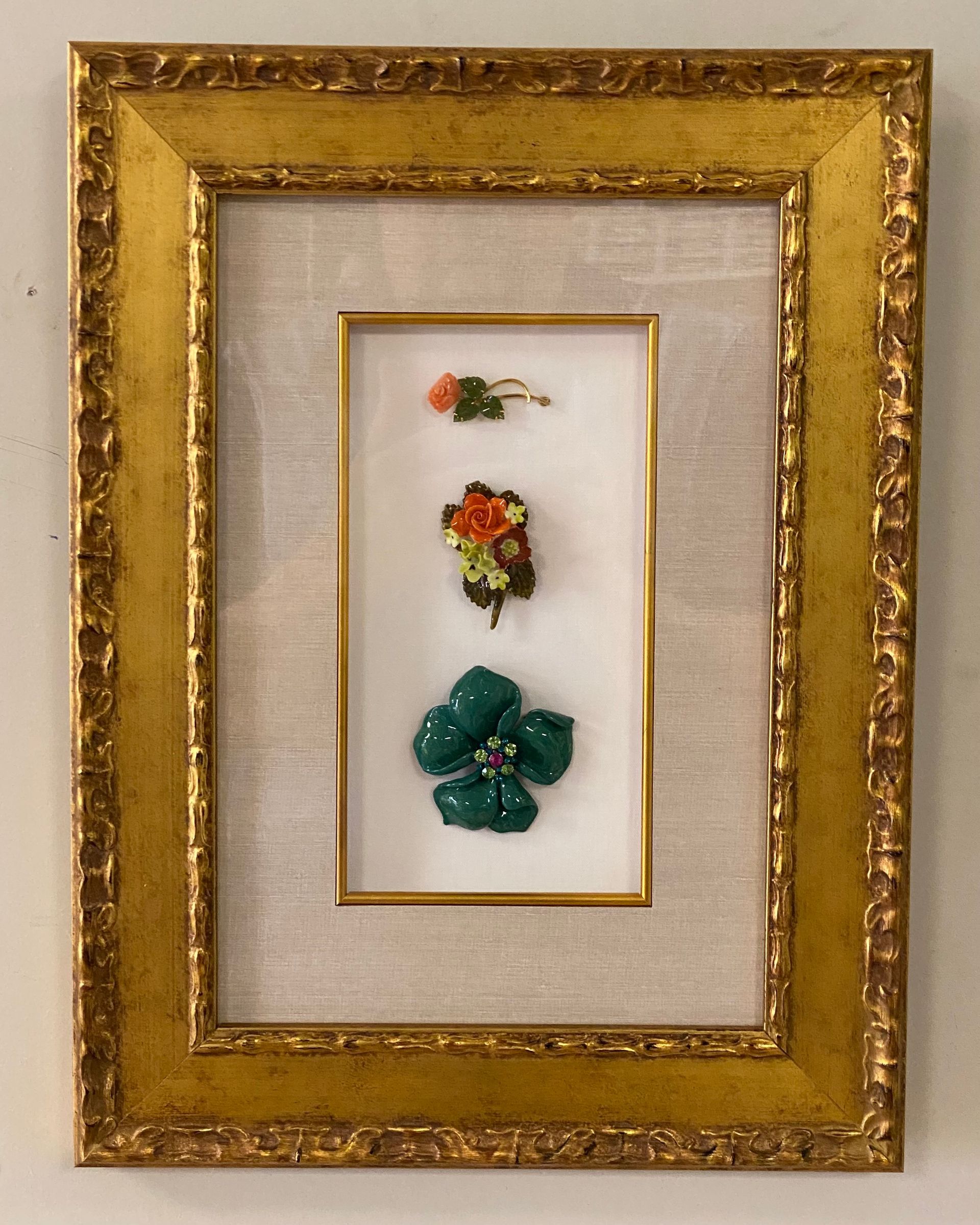 A framed display of three decorative flower brooches: a small orange flower, a colorful cluster, and a large green jewel.