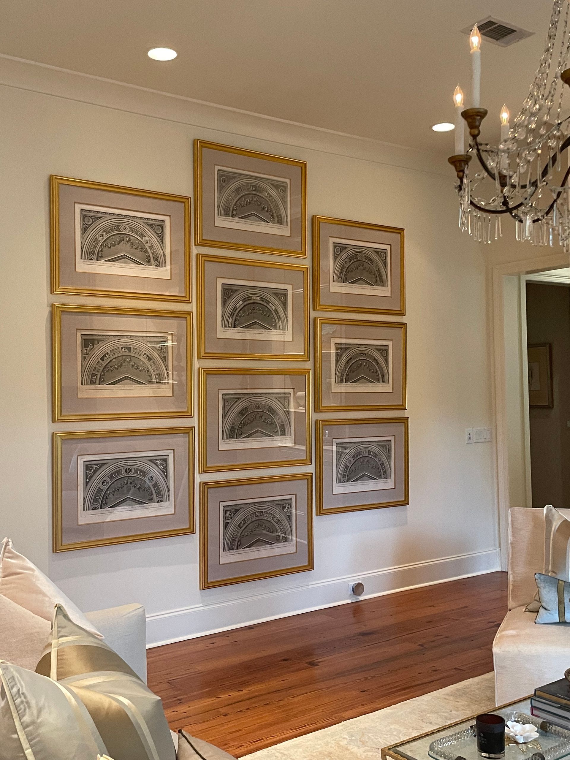 A wall of framed black and white art hangs in a room. The frames have gold trim. A chandelier hangs nearby.