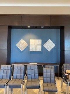 Three geometric wall art pieces above a table in front of a blue wall. Gray chairs sit in a row, and the background is brown.