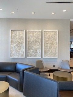 Three framed textured art pieces hang above a dark leather sectional sofa in a modern waiting area.