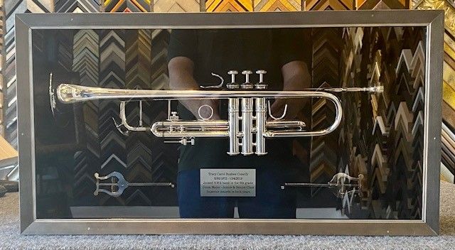 A silver trumpet displayed in a shadow box frame, with a name plate, set against a background of various frame samples.