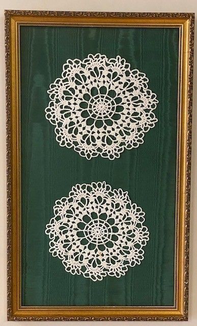 Framed artwork featuring two white lace doilies against a dark green background, within a gold ornate frame.