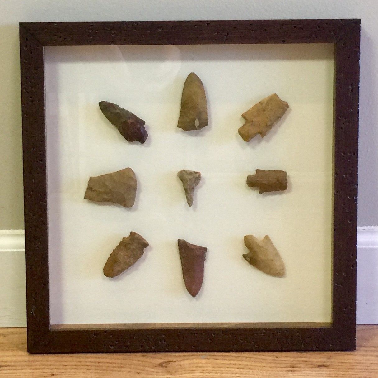 A framed display of nine arrowheads, arranged on a cream-colored background. 