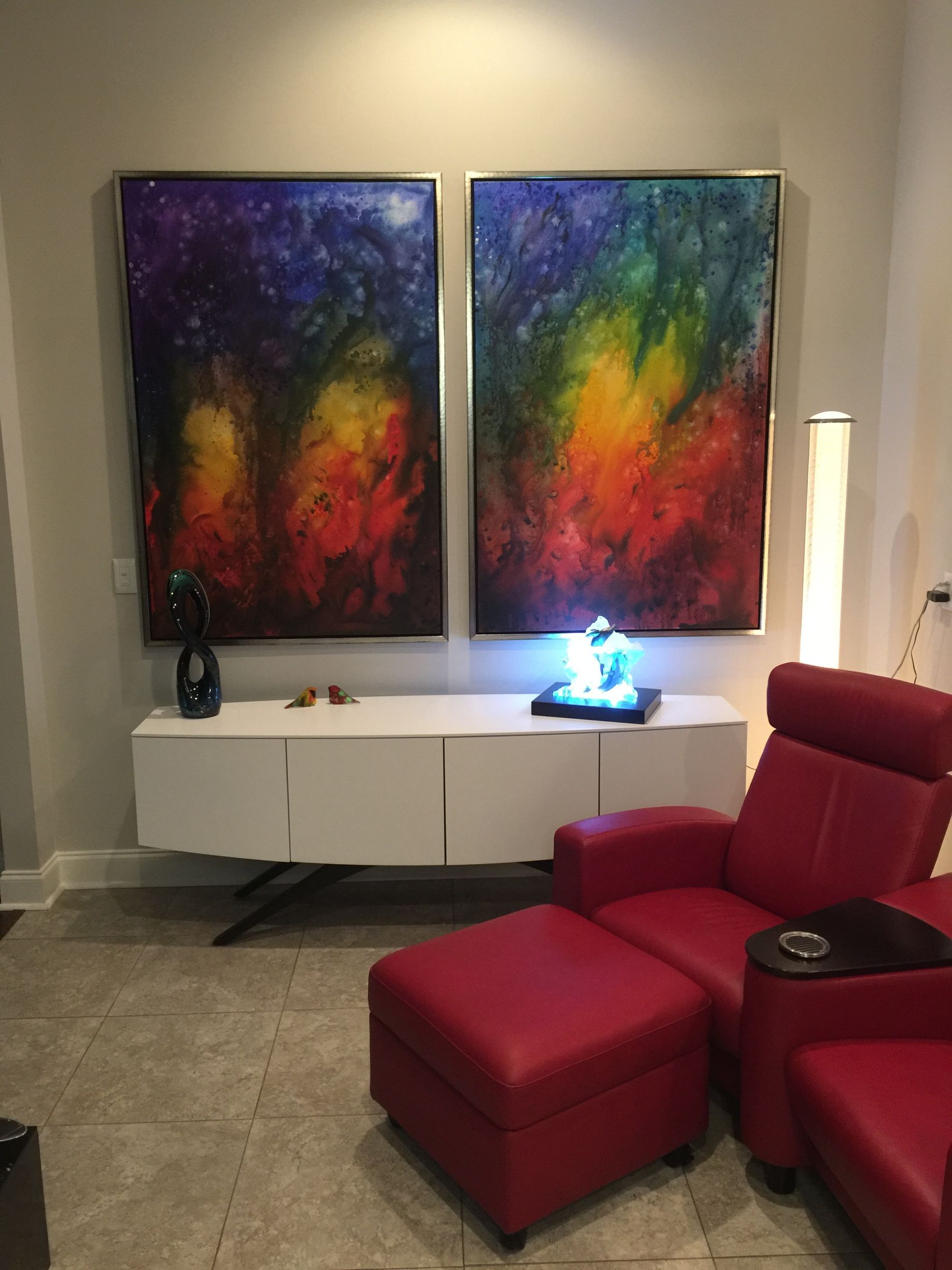 Two colorful abstract paintings hang above a white cabinet. A red recliner and ottoman sit to the right.