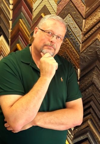 Man with glasses and a green polo shirt, posing with his hand on his chin in a frame shop.