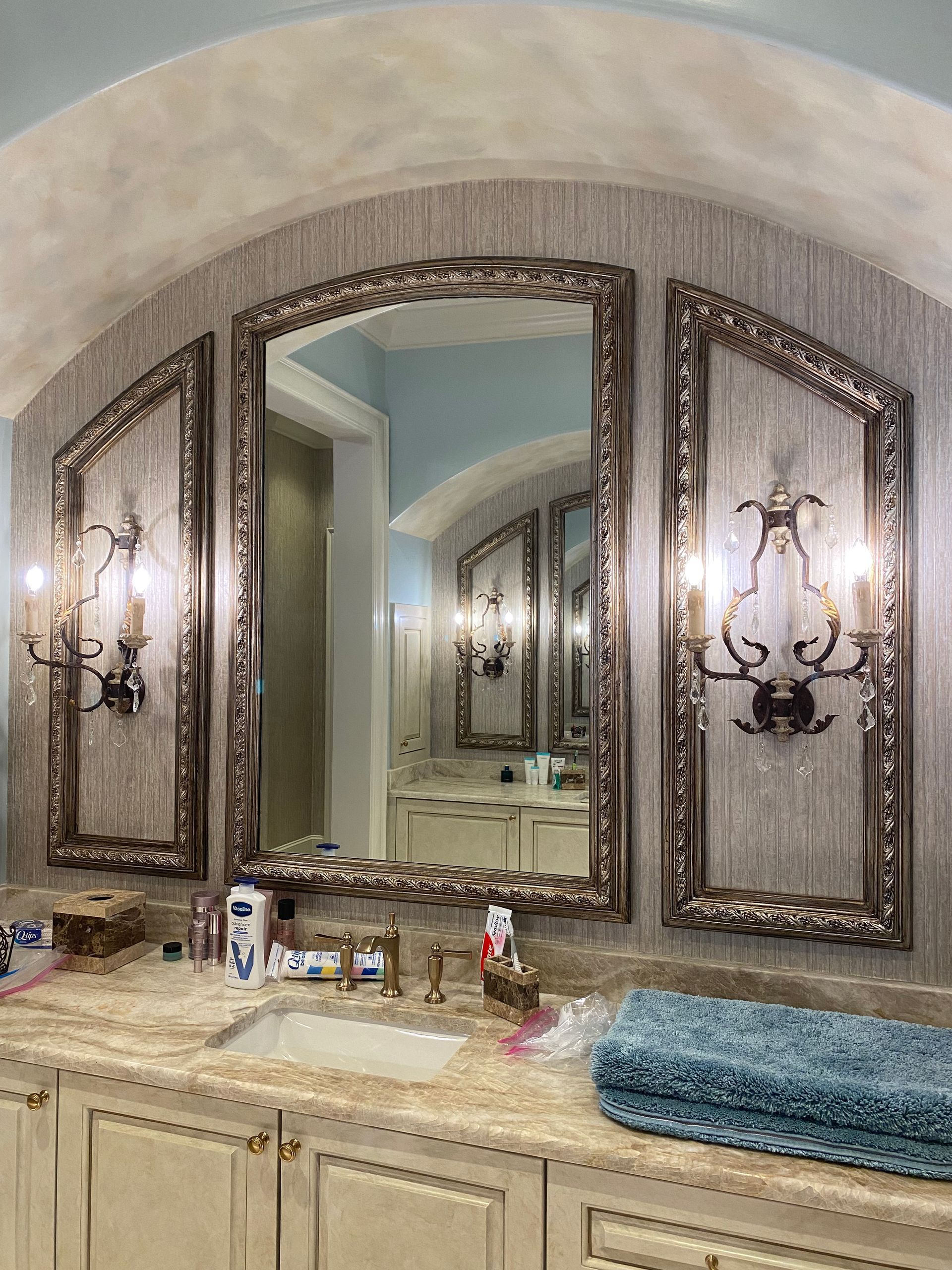 Bathroom with three ornate framed mirrors and sconces above a countertop and sink. The walls are a textured silver-gray.