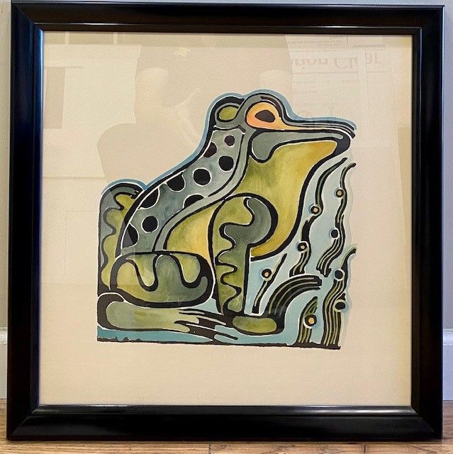 Framed watercolor painting of a stylized frog in shades of green, blue, and yellow. The frog sits against a pale background.