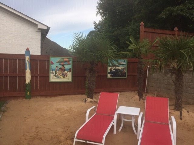 Backyard beach scene with red lounge chairs, sand, palm trees, and artwork on a brown fence.