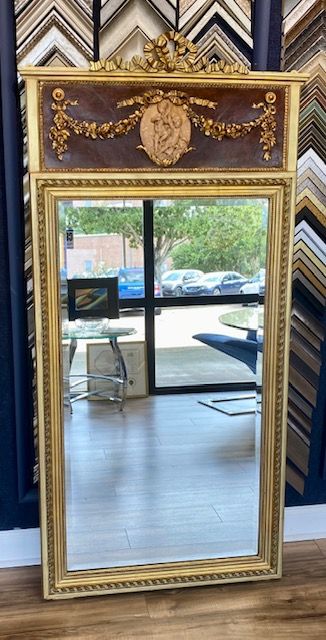 Tall, ornate, gold-framed mirror with a decorated top panel. 