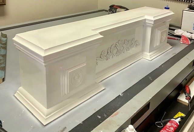 White wooden decorative mantelpiece on a gray surface, showcasing ornate carvings. It sits in an indoor setting.