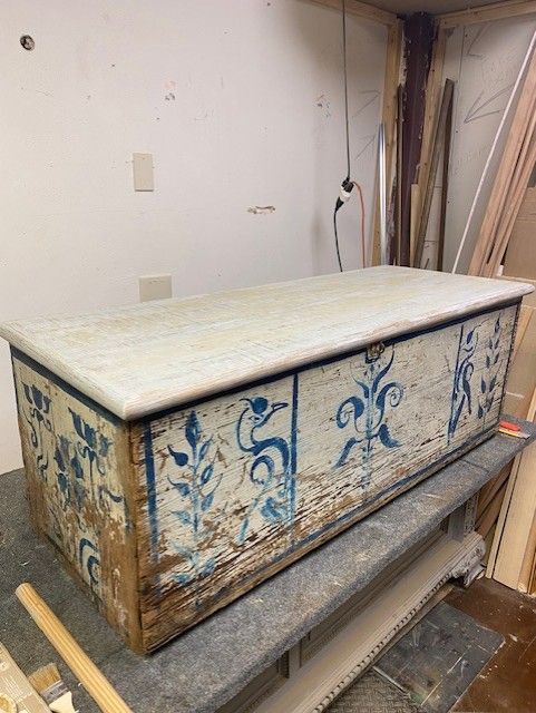 A weathered wooden trunk with faded blue floral painted designs, sitting on a gray work surface in a workshop.