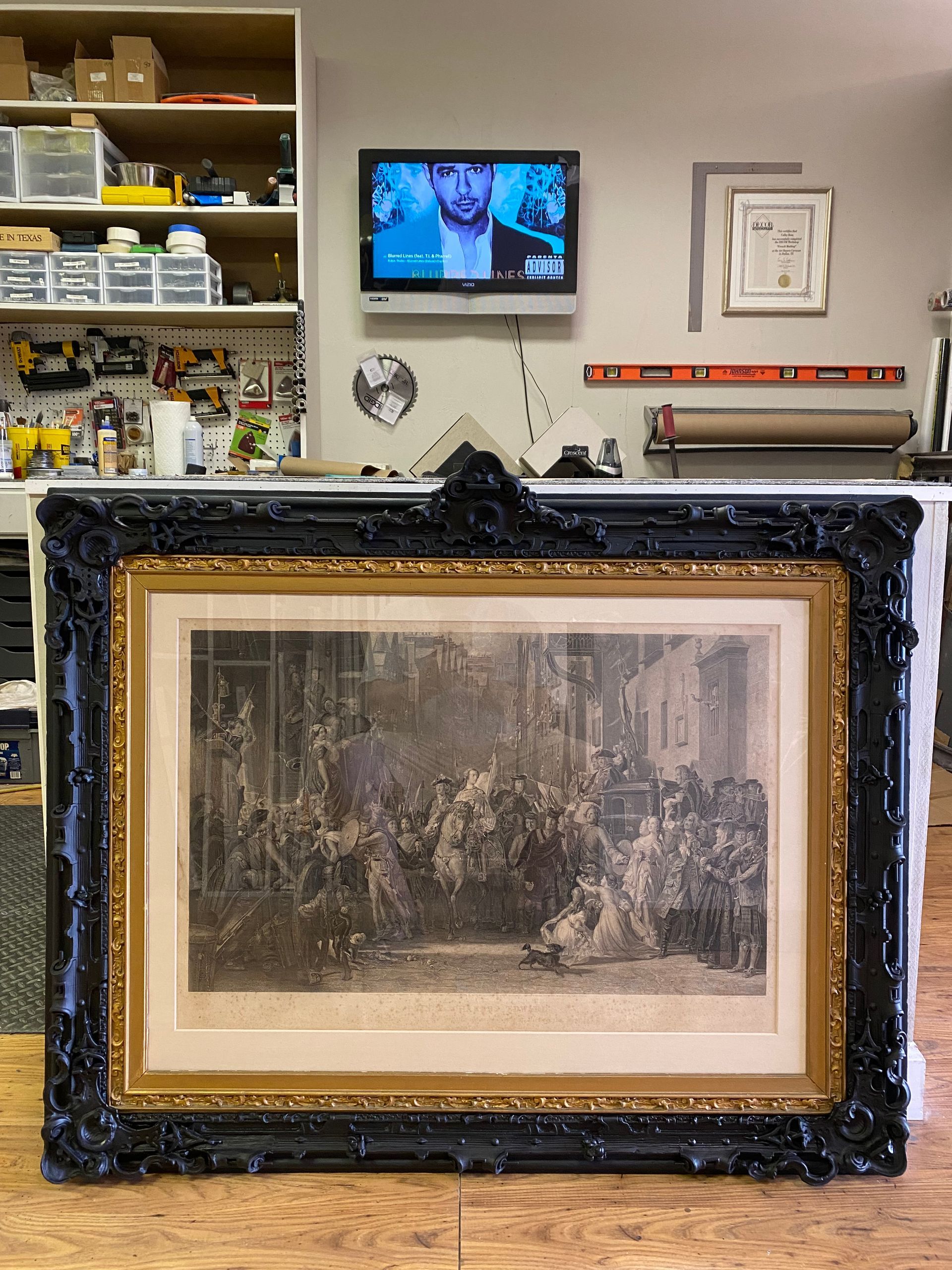 Large, ornate framed artwork of a detailed black-and-white scene, leaning against a work surface.