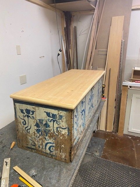 An old chest with peeling blue floral paint and a new light wood top in a workshop.