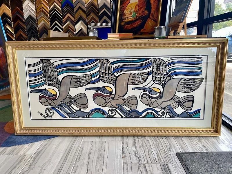 Artwork of three pelicans flying over stylized waves, framed in gold. The background includes a framing store.