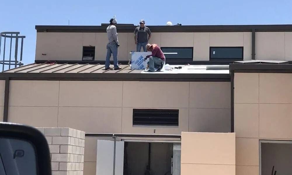 Three workers on a flat rooftop inspecting or repairing the roof of a beige building.