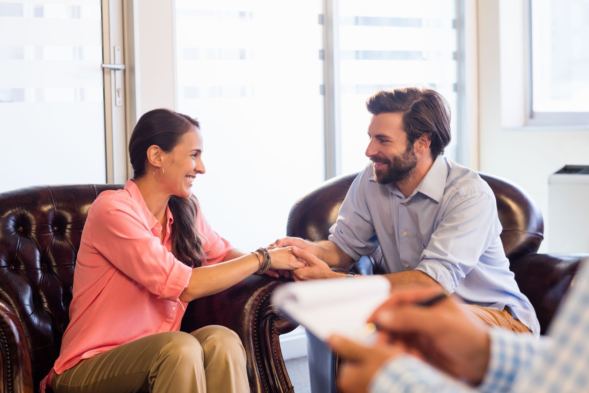 Two people smiling and shaking hands in a bright office, with a third person blurred in the foreground