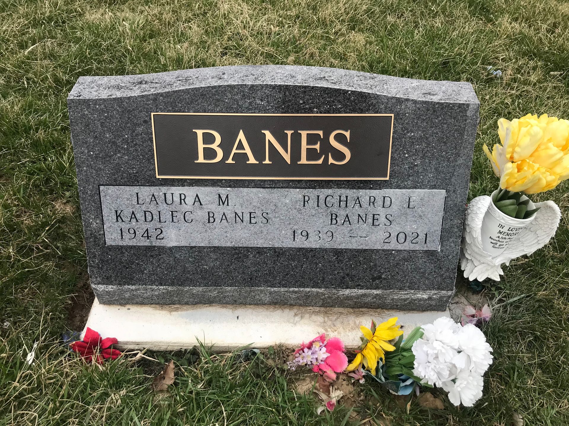 A gravestone for Banes is surrounded by flowers in a cemetery