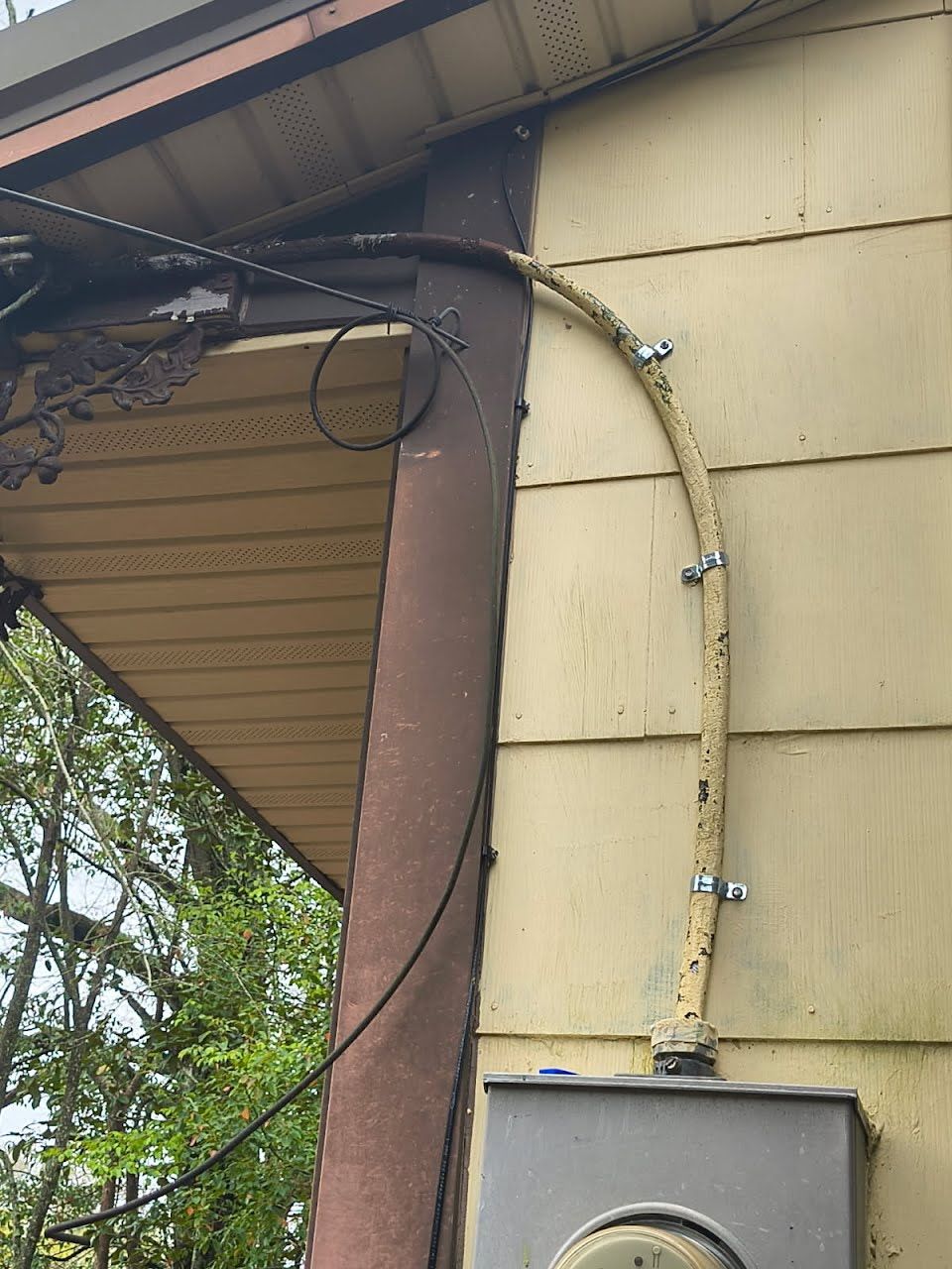 A beige electrical conduit runs down the side of a house, secured by metal brackets to an electric meter base.