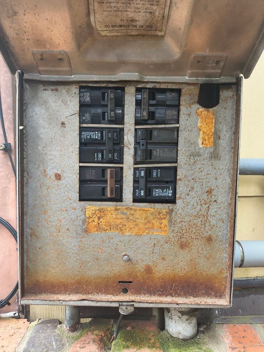 An open, weathered, and rusty outdoor electrical breaker box with six installed circuit breakers.
