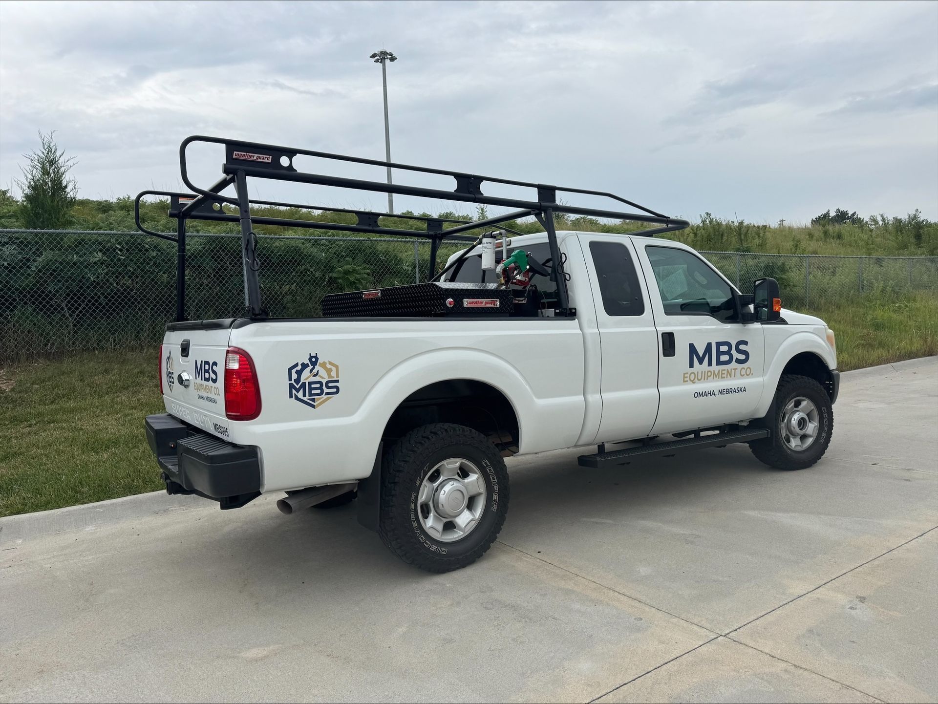 White utility pickup truck with ladder rack parked on a concrete road beside a grassy field.