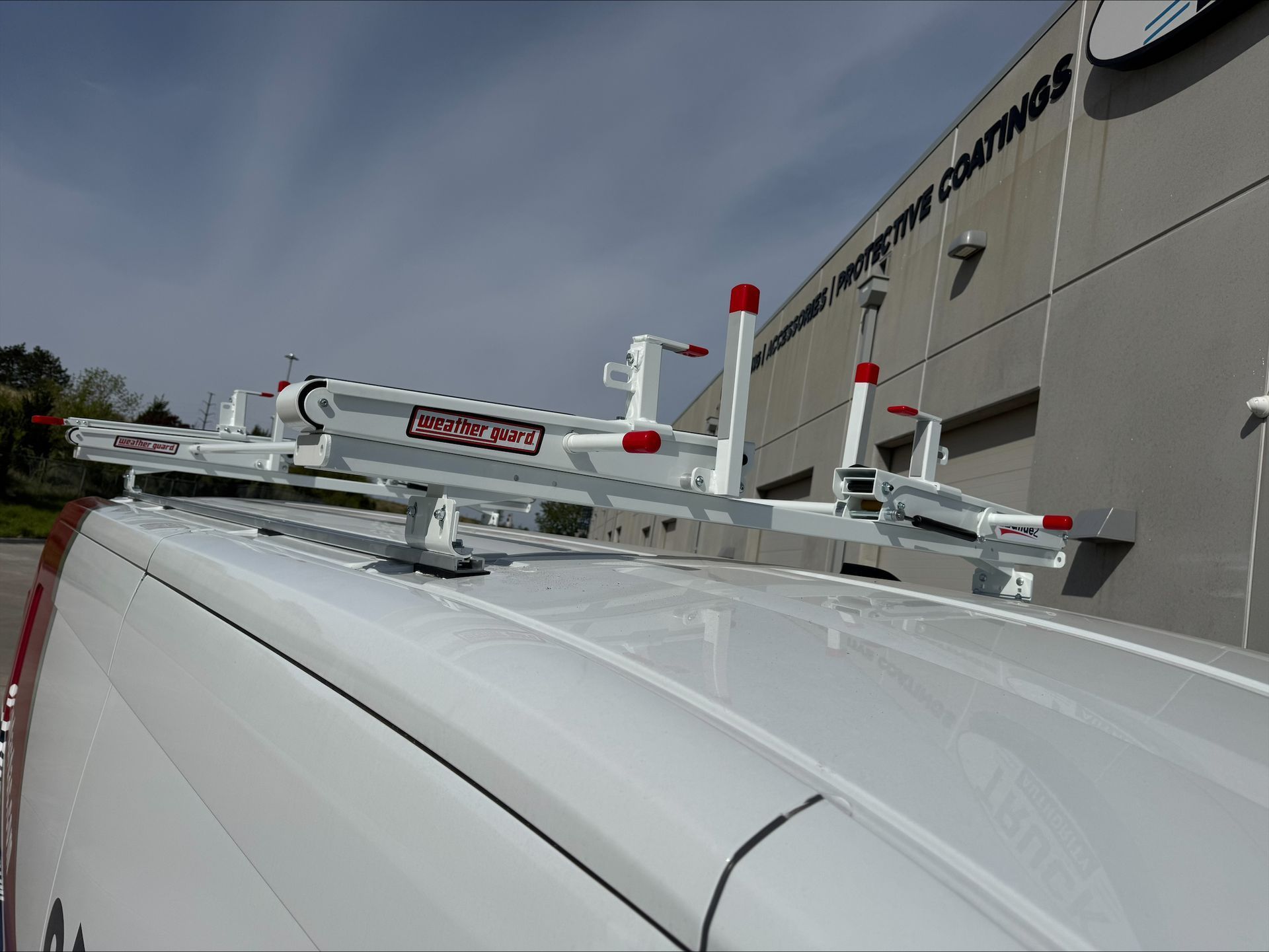 White service van roof with mounted ladder racks and equipment beside a commercial building.