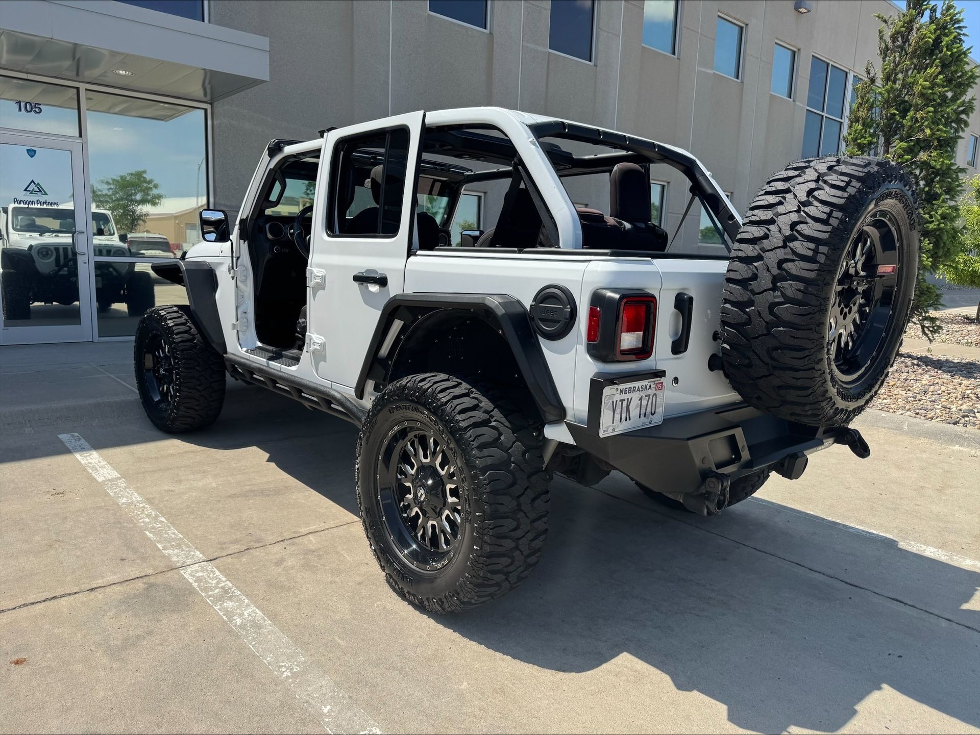 White Jeep Wrangler with doors removed, parked outside a building on a sunny day.