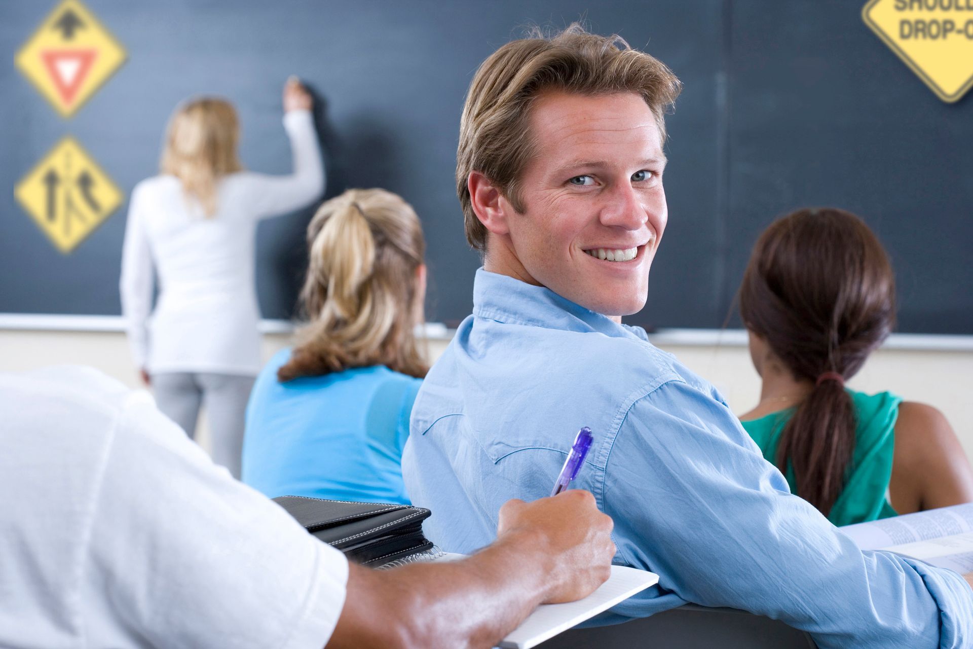 Man smiles in a classroom; students and chalkboard with road signs in the background.