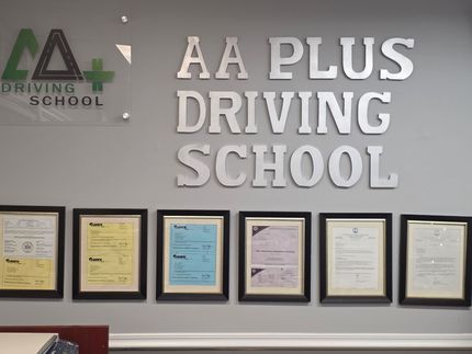 AA Plus Driving School sign with framed certificates on a grey wall.