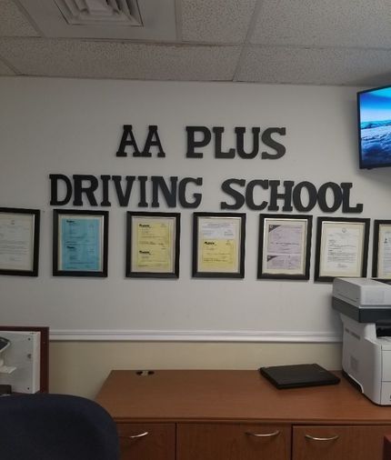 AA Plus Driving School sign above framed documents on a white wall in an office.