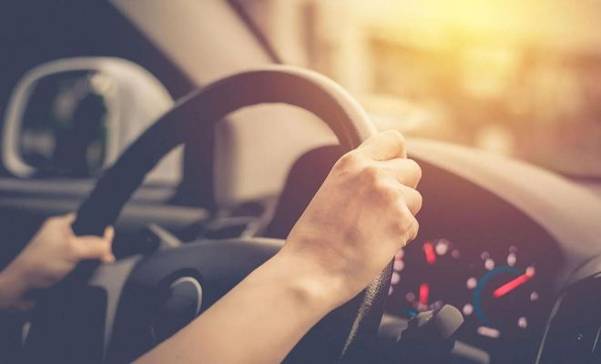 Person's hands gripping a car steering wheel with a dashboard visible, bright sunlight in background.