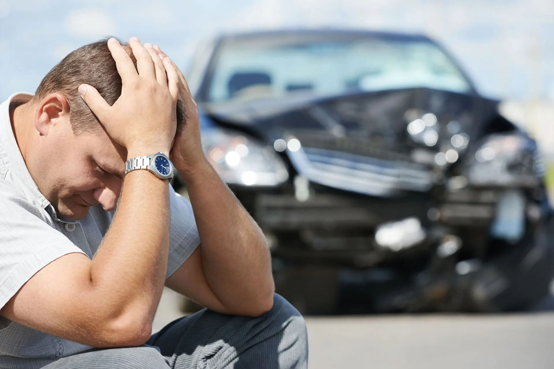 Man with head in hands sits in front of a badly damaged black car.