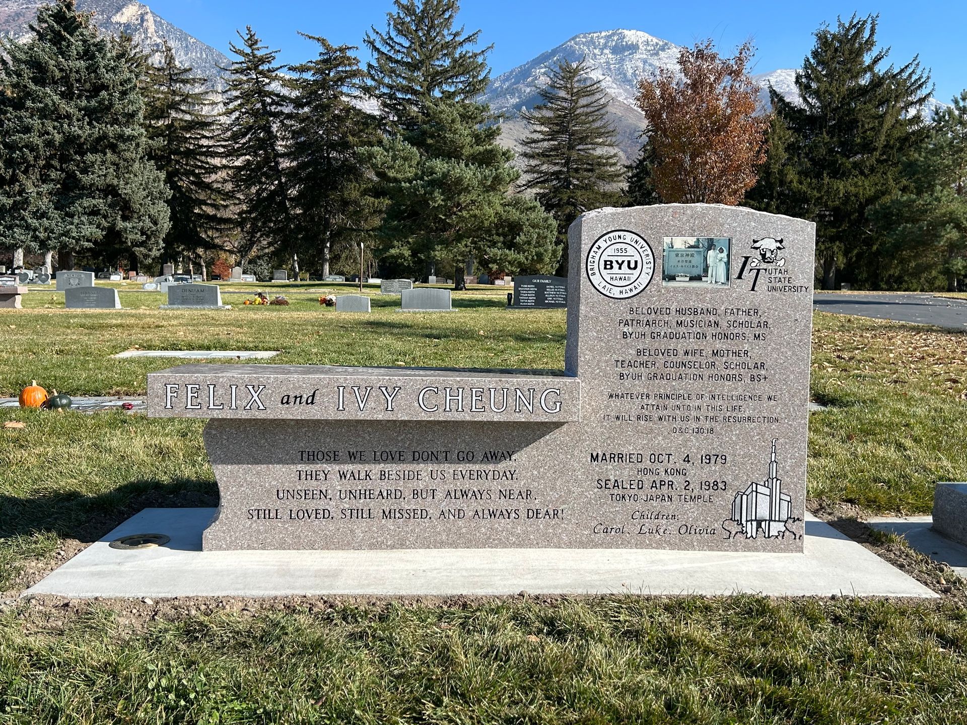 Grave marker with bench in cemetery, mountains in background. Gray stone, inscribed with text.