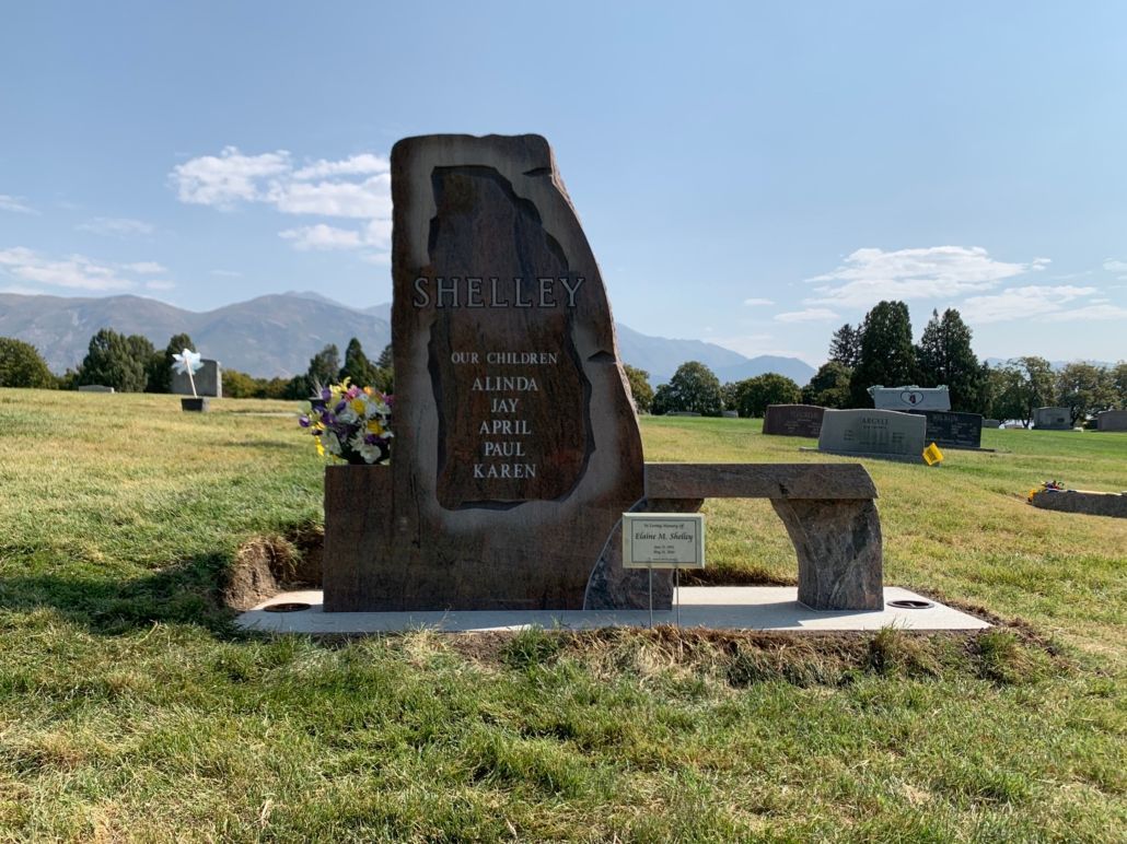Grave marker with bench, Shelley inscribed. Yellow flowers sit at base. Green grass, blue sky, mountains.
