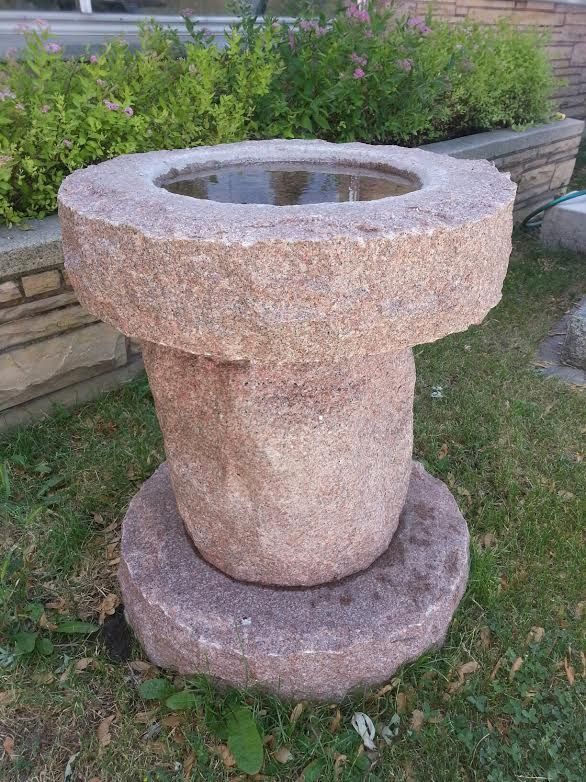 Stone bird bath, pinkish-gray color, round basin atop a pedestal. Green shrubbery in the background.
