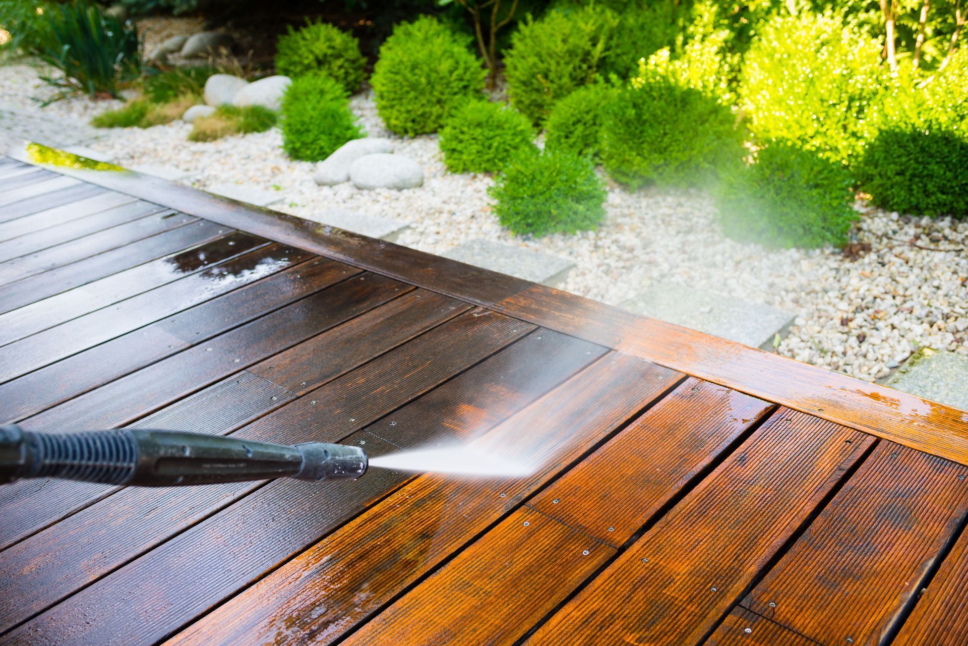 A person pressure washing a wooden deck, cleaning it; water spraying, some boards cleaned, others dirty.