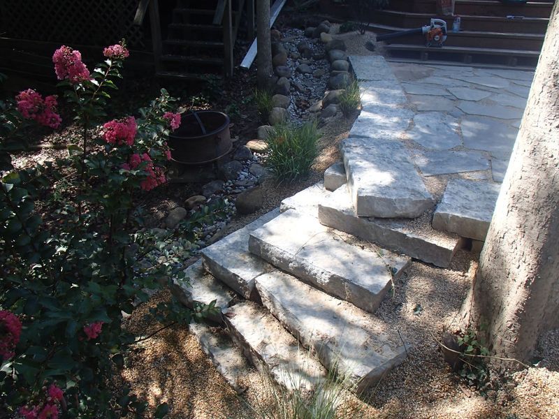 Stone steps and pathway leading up a hill, next to a tree and garden with pink flowers.