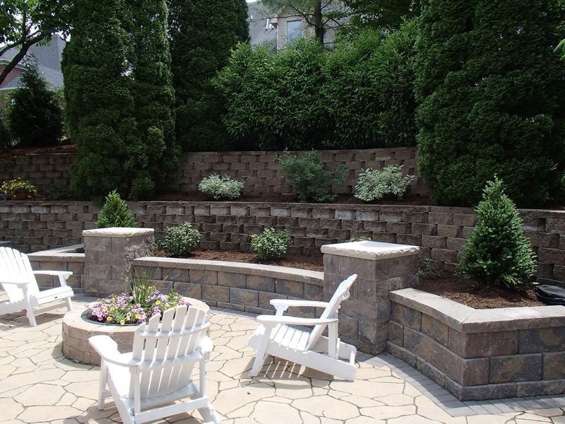 Patio with white chairs, fire pit, and retaining walls with greenery.