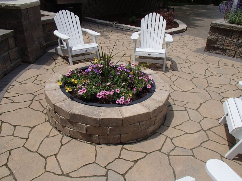 Two white Adirondack chairs face a circular flower bed on a stone patio.