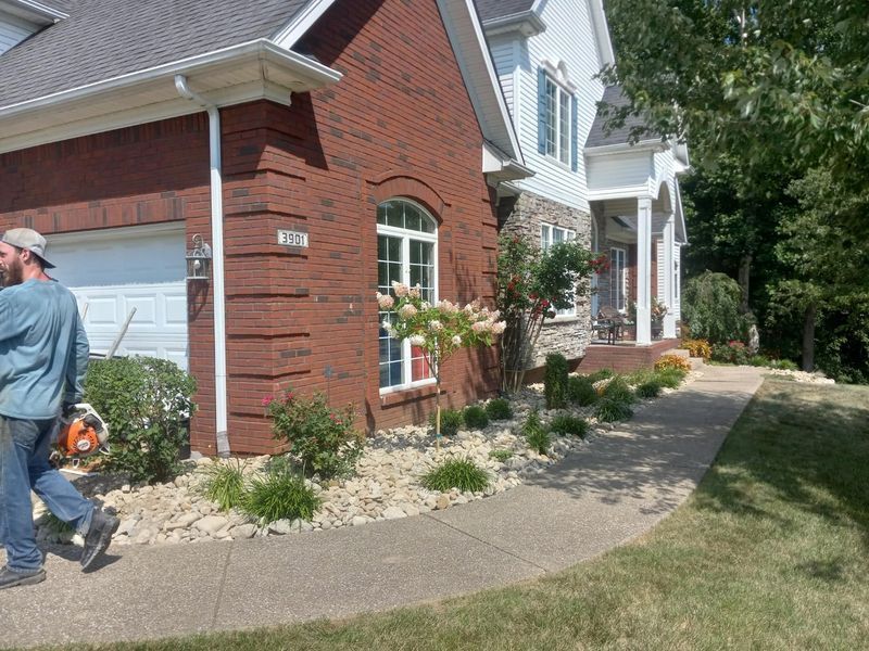 Man with chainsaw near red brick house with landscaping along a sidewalk.