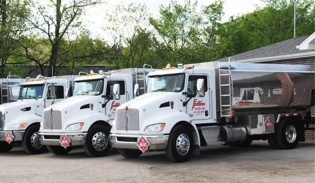 Three white dump trucks parked in a row on a gravel lot, with trees and a canopy in the background