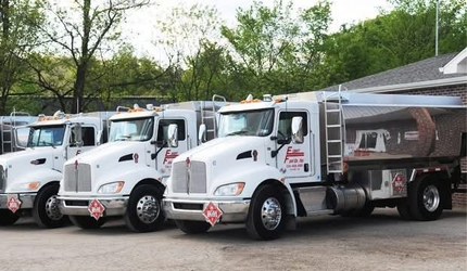 Three white dump trucks parked in a row on a gravel lot, with trees and a canopy in the background