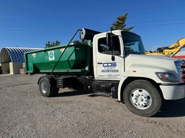 A white CDS roll-off dump truck with a green container parked in a gravel lot under a clear blue sky.
